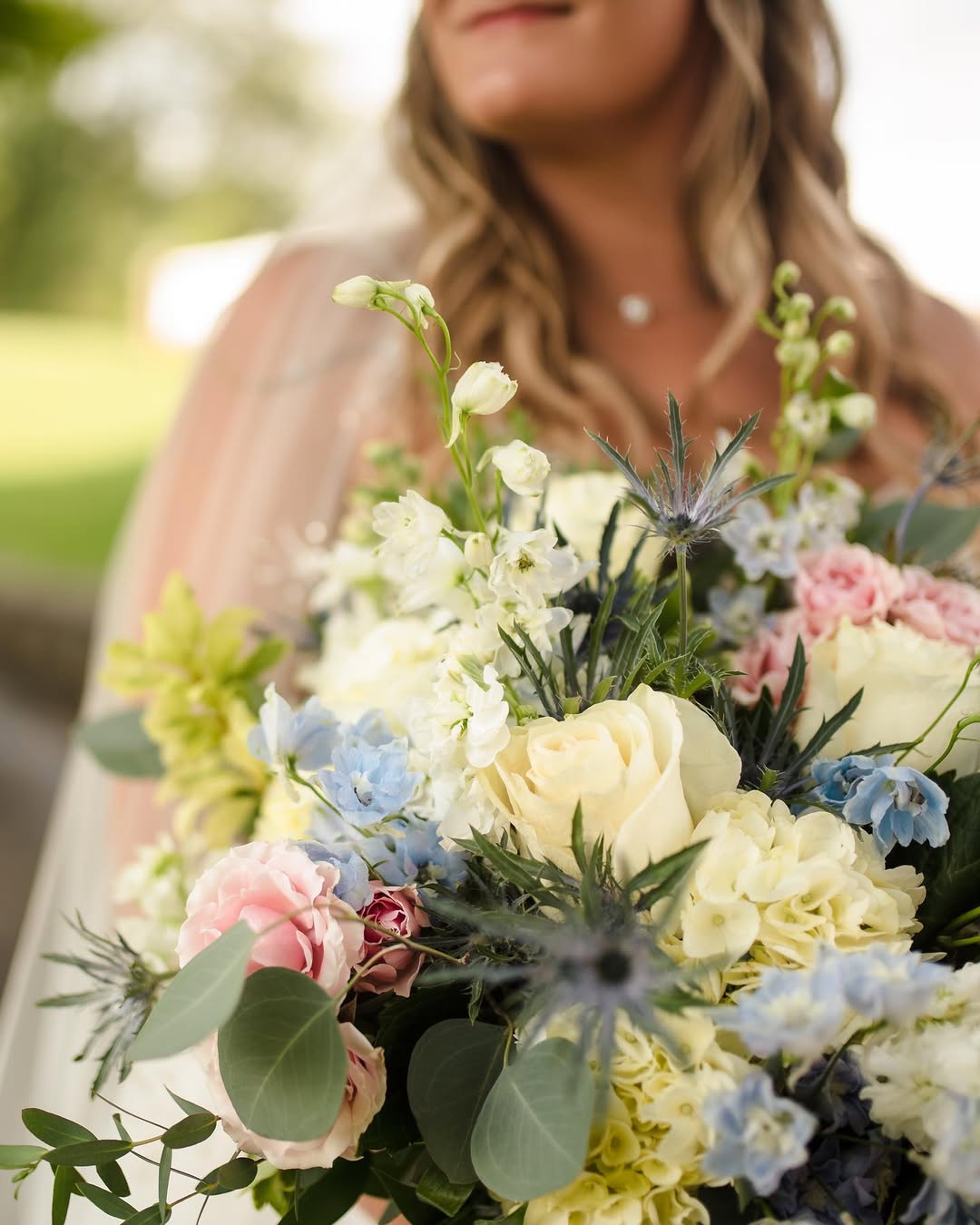 Bridal bouquet of ivory roses, blush carnations, blue delphinium and thistle held by a bride