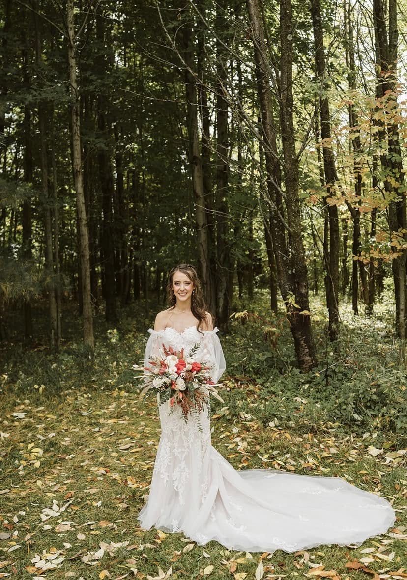 Bride in a lace gown holding a burgundy and ivory bouquet in a wooded clearing