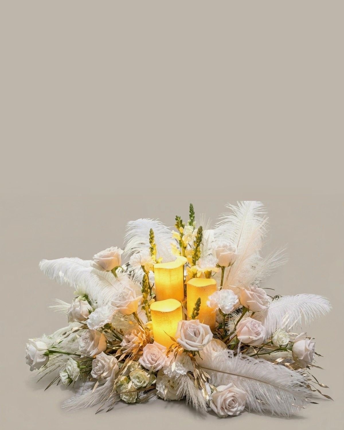 Low centrepiece in white and cream with three lit pillar candles, ivory roses, ostrich feathers, pampas and gilded leaves
