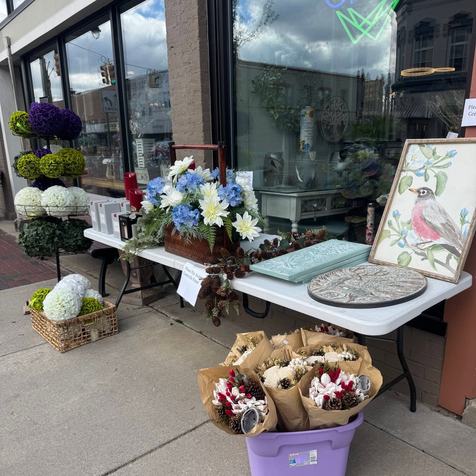 The front of Greenville Floral on South Lafayette Street in Greenville, Michigan: the shop's window bay and brick pillars, with a table of arrangements and gifts set out on the sidewalk in front of it.