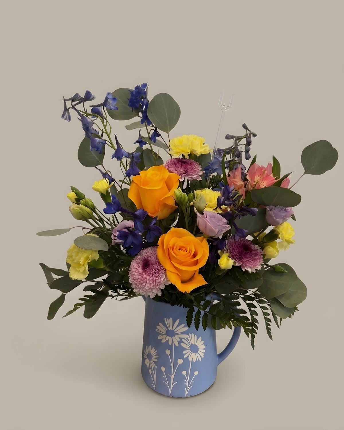 Orange roses, yellow blooms and purple accents in a blue floral pitcher