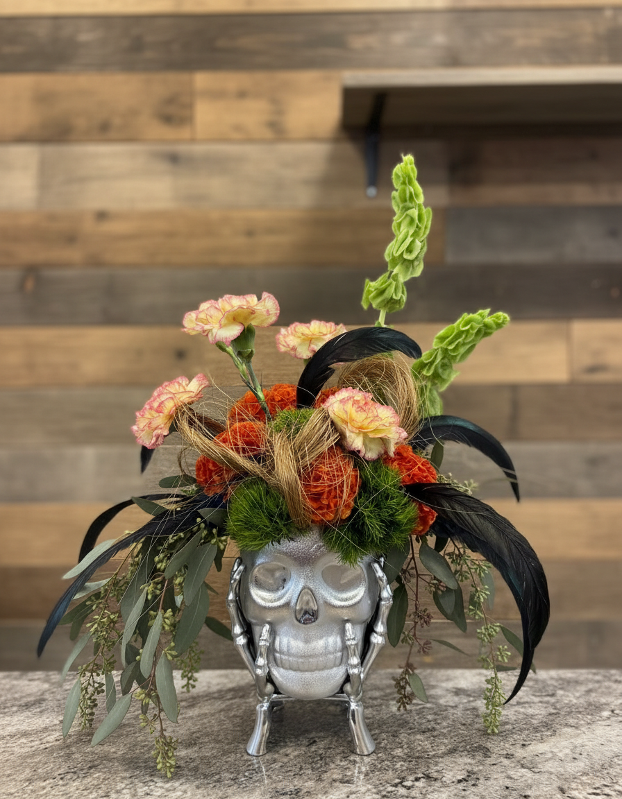 Orange carnations, bells of Ireland, eucalyptus and black feathers in a silver skull vase