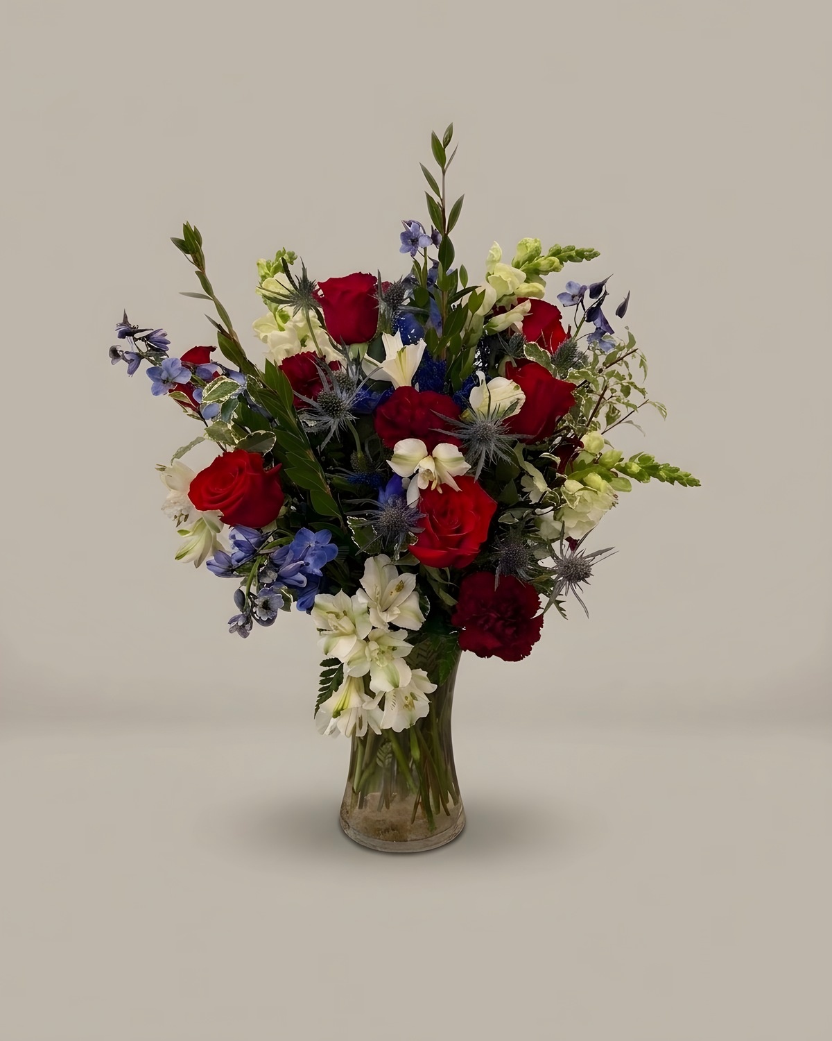 Red carnations, white blooms and blue delphinium in a clear glass vase